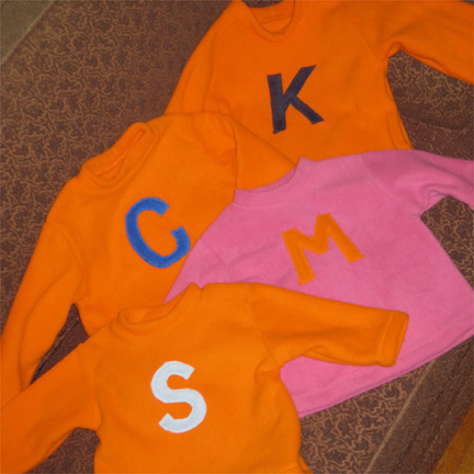 KCMSshirts.jpg - A family of four, with color choices to tie together the three boys (orange shirts with different shades of blue letters) with their sister (pink shirt with an orange letter)